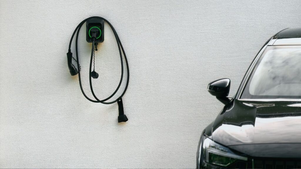 Een zwartkleurige wallbox die bevestigd is op een witte muur. Rechts hiervan staat een zwarte auto geparkeerd.
