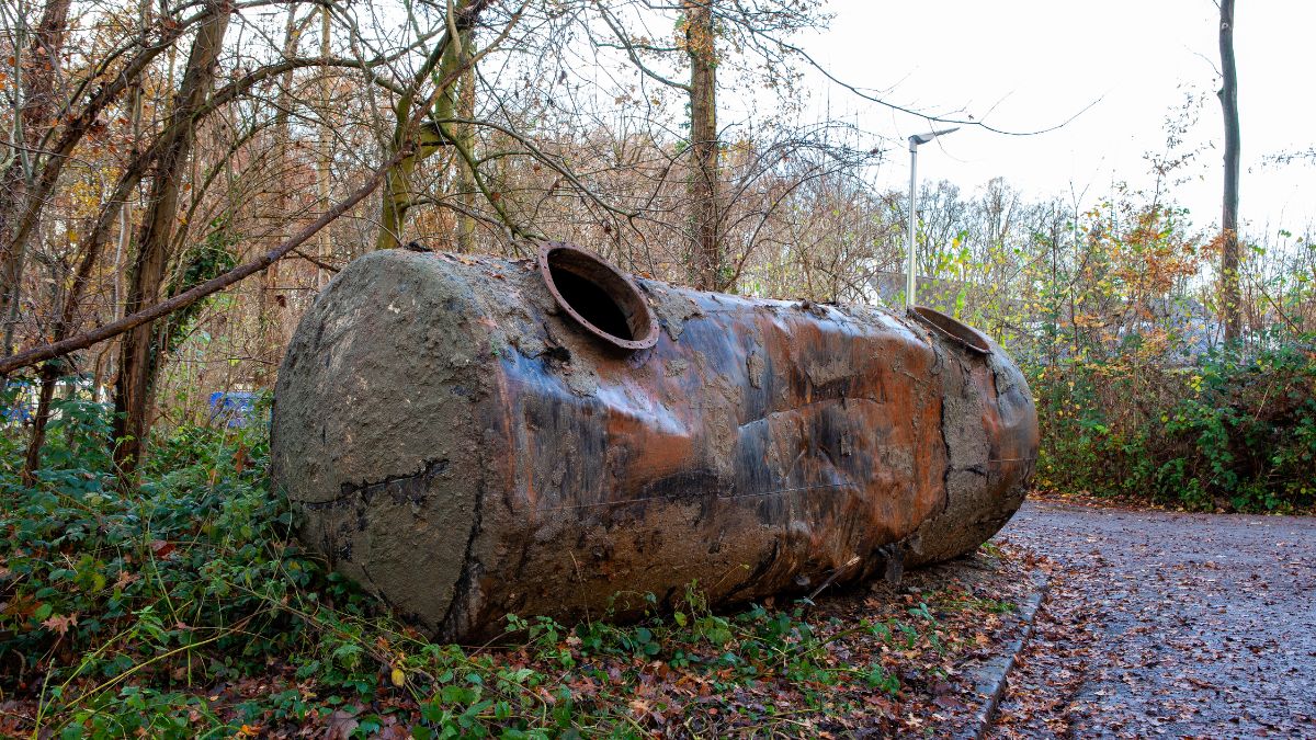 Een oude lege mazouttank langs de kant van een weg. De tank ligt naast een stel jonge bomen.