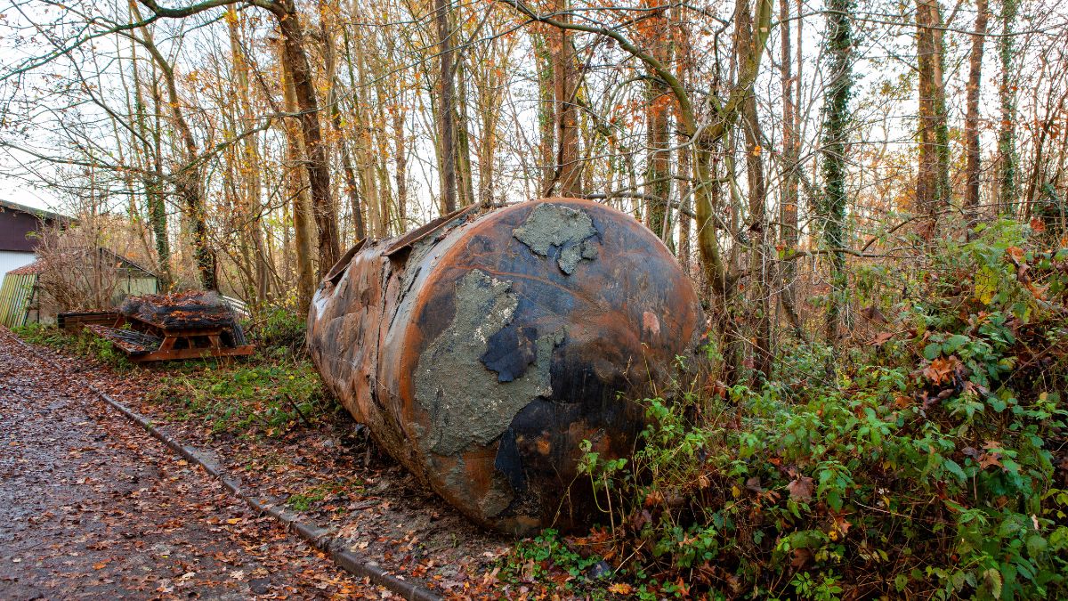 Een oude, uitgegraven mazouttank met roestsporen. De tank ligt langs de kant van een pad door een bos.