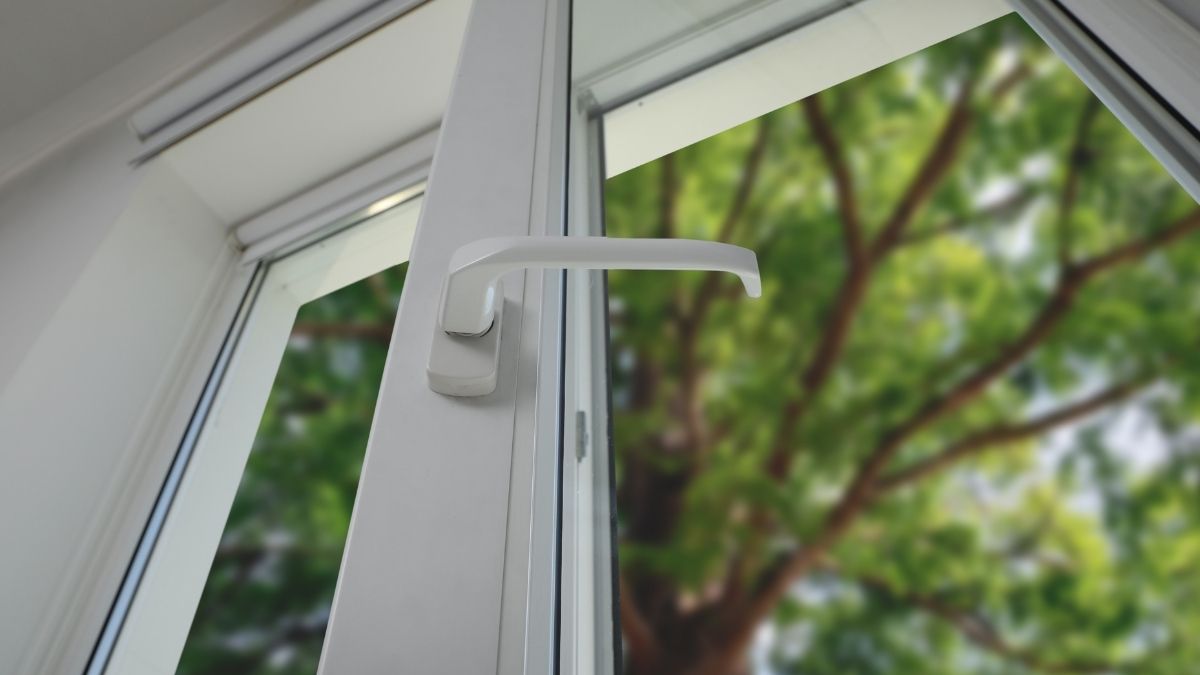Een raam in wit pvc. Het raam is deels geopend, en de klink wijst naar rechts. Achter het raam staat een grote boom met bladeren.