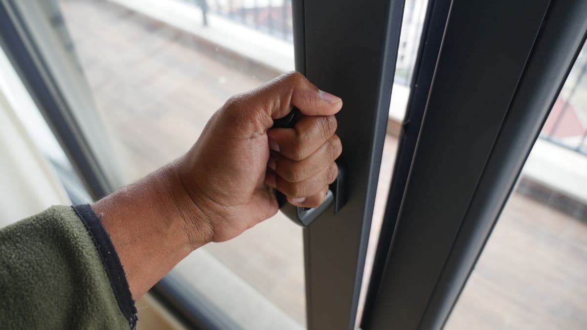 Een hand opent een kantelraam of valraam. Het raam heeft zwarte kozijnen en geeft uit op een terras. De hand komt uit een groene mouw in fleecestof, afgewerkt met een zwarte boord.