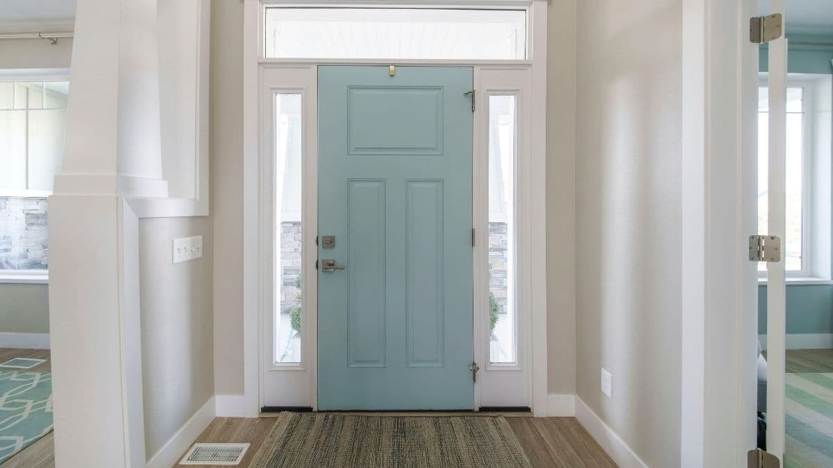 Een moderne inkomhal met lichtgrijze muren, parketvloer en een lichtblauwe voordeur. Boven, links en rechts van de voordeur zitten smalle ramen.