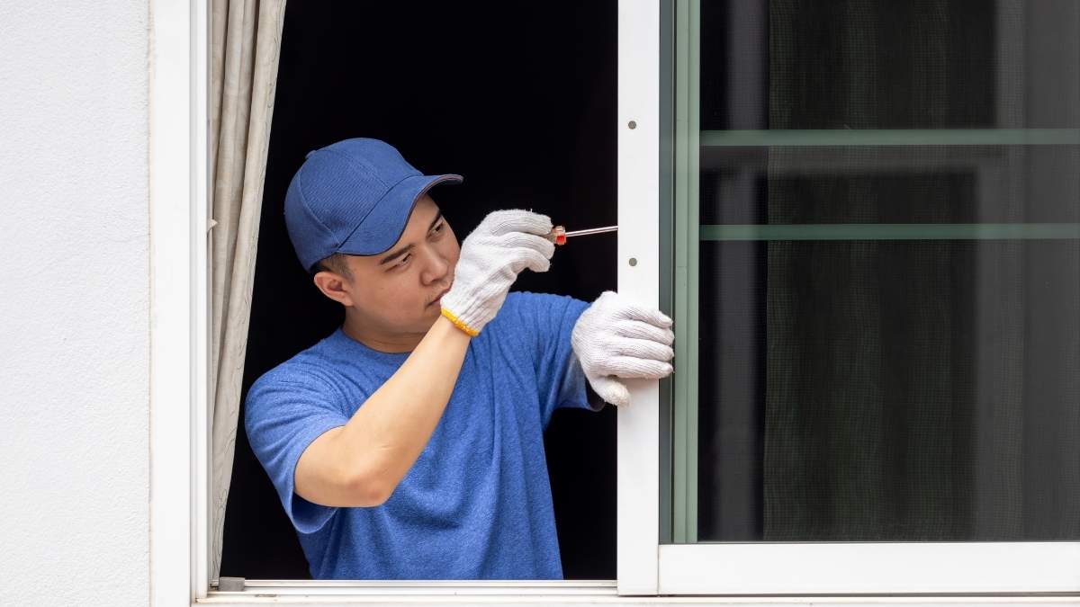 Een raamspecialist in blauw t-shirt, blauwe pet en met witte werkhandschoenen werkt aan het hang- en sluitwerk van een wit raam in pvc. Het raam vertoont wat gebruikssporen. De expert gebruikt een schroevendraaier om de klink van het raam af te stellen.