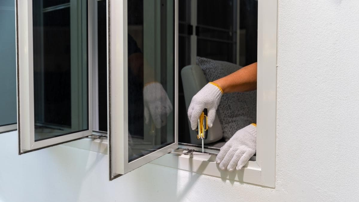 Een raam met drie luiken en witte raamkozijnen in een witte gevel. Uit het dichtstbijzijnde raam komen twee handen in witte werkhandschoenen. De handen gebruiken een kleine schroevendraaier om de nieuwe ramen in de gevel te bevestigen. 
