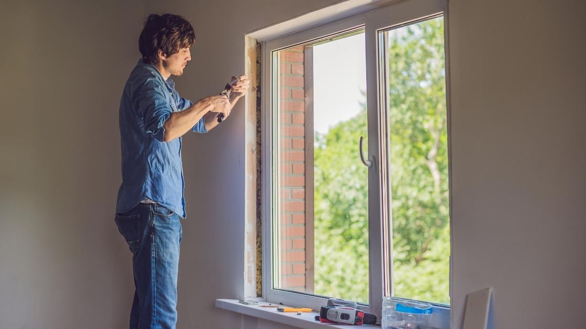 Een raamspecialist in jeansbroek en blauw hemd werkt de binnenkant van een nieuw raam in pvc af. Op de vensterbank liggen verschillende gereedschappen.