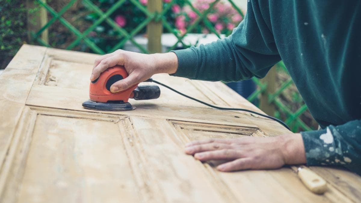 Een schrijnwerker in groene trui schuurt een houten deur met een oranje handschuurmachine. Hij zit in openlucht, voor een groen hek.