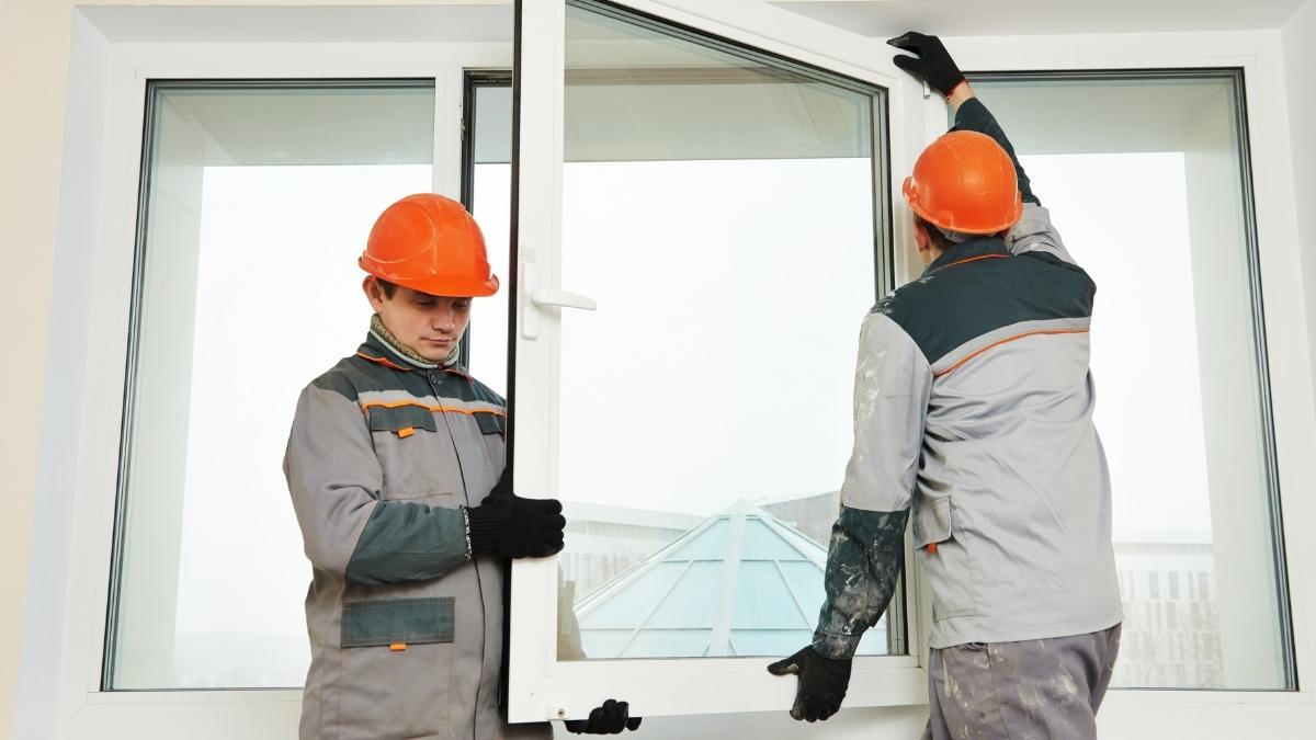 Twee ramenzetters met oranje helmen en grijze werkkledij plaatsen een nieuw pvc raamvleugel in een pvc raamkozijn. In het glas is de reflectie van een blauw puntdak zichtbaar. De linkse ramenzetter kijkt schuin naar beneden. De rechtse ramenzetter kijkt naar de rechterbovenhoek van de raamvleugel.