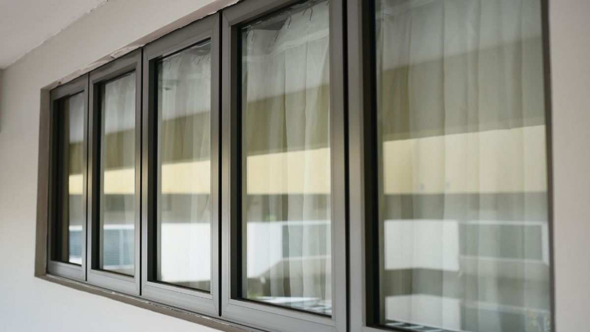 Een gebroken witte gevel, voorzien van 5 ramen op een rij. De ramen zijn voorzien van licht getint zonwerend glas en zitten in grijze kunststof raamkozijnen. Achter de ramen hangt een wit gordijn.