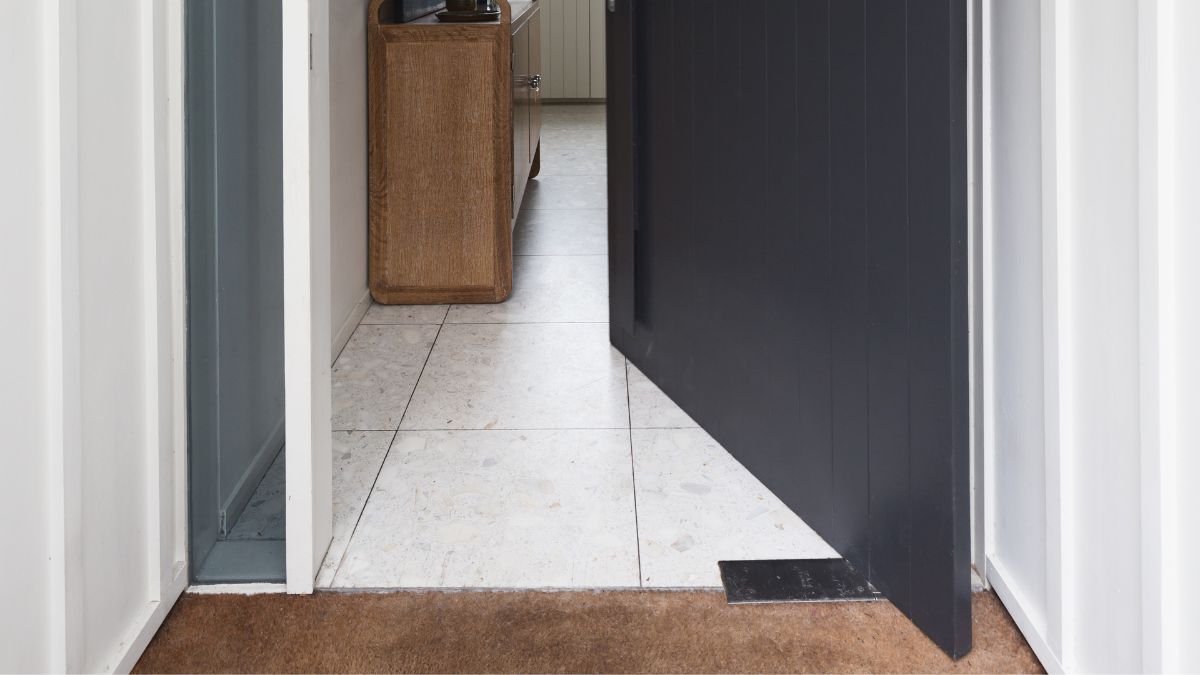 Detail van de onderzijde van een excentrische pivotdeur in zwart hout. De deur is geopend. Achter de deur ligt een ruimte met tegels, waarin een lage houten kast staat.