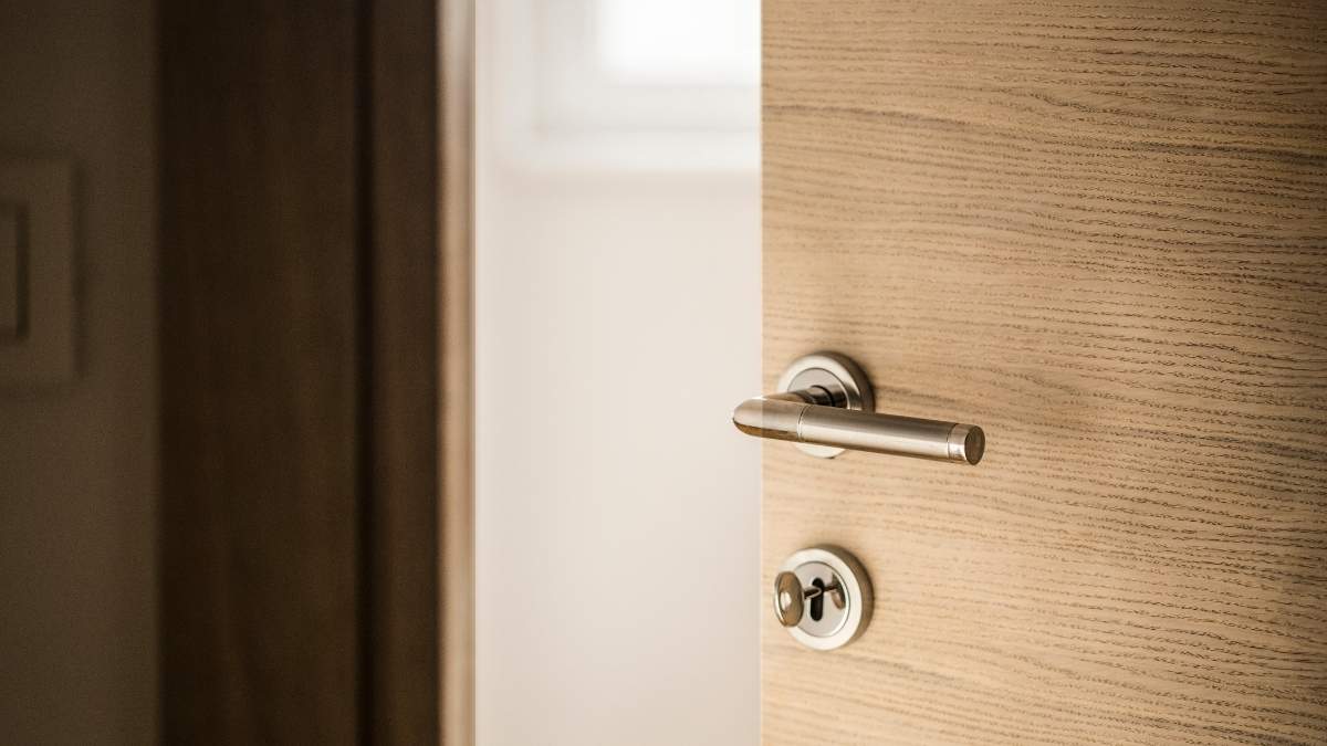 Detail van een geopende houten deur met slot en metalen klink. De deur zit in een houten deurkader. Achter de deur ligt een witte kamer met raam.