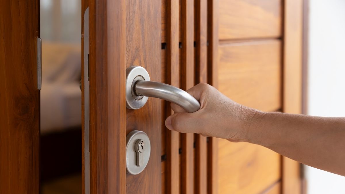 Een houten buitendeur van het type opdekdeur. Een hand houdt de metalen klink van de voordeur vast. De deur  is half geopend.