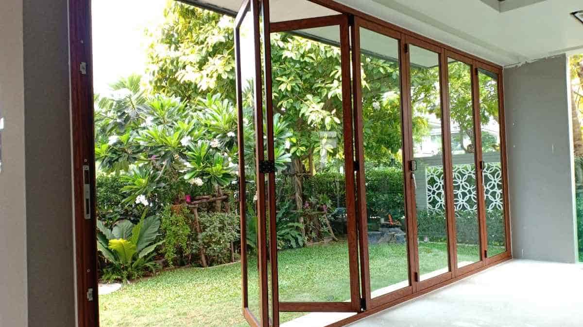 Een houten vouwraam met 6 panelen. Het vouwraam geeft uit op een gazon, omringd met tropische planten.
