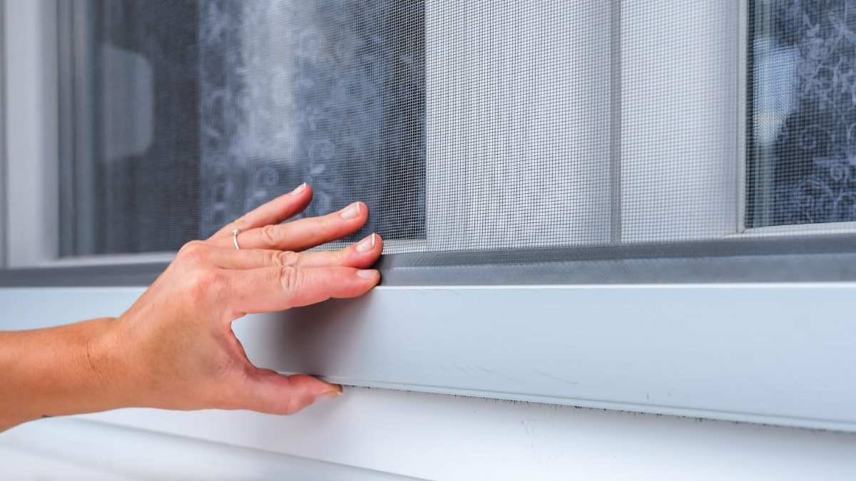 Een vrouwenhand met ring om de ringvinger houdt de onderste lat van het profiel van donkergrijze screens vast. Achter de screens ligt een raam met witte kozijnen.