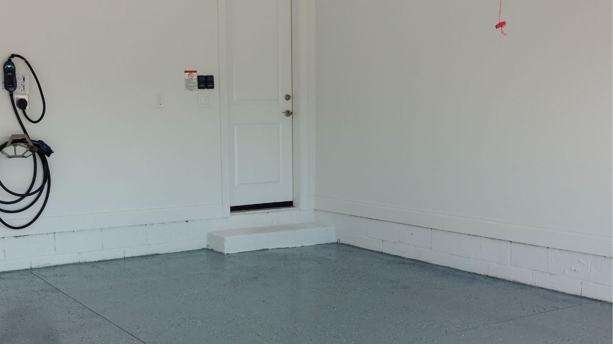 Een garage met witte muren en een grijze vloer. Links aan de muur hangt een laadpaal in wallbox model. Rechts naast de laadpaal, in dezelfde muur, zit een isolerende binnendeur die de garage met de rest van de woning verbindt.