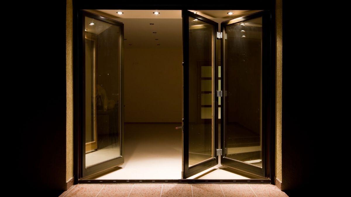 Een zwarte vouwdeur met grote ramen tussen een lege kamer en een terras met rode tegels. De deur bestaat uit 2 delen: links een geopende scharnierdeur, rechts 2 half geopende vouwpanelen.