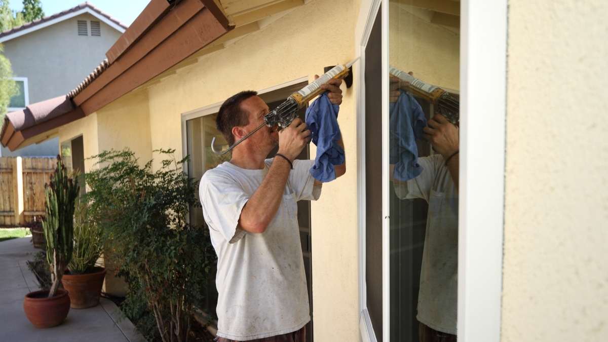 Een doe-het-zelver in een vuil wit t-shirt gebruikt een kitpistool om de buitenkant van witte ramen af te kitten. In de witte raamkozijnen zit spiegelend glas. De man houdt, naast het kitpsitool, een blauwe pluisvrije doek vast. De ramen zitten in een gele gevel onder een bruin dak. Tegen en voor de gevel staan verschillende terracotta potten met planten.
