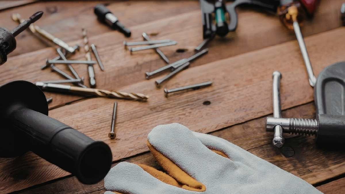 Gereedschap op een ruwe houten tafel. Er liggen onder meer nagels, schroeven, een lijmklem en een grijs met gele werkhandschoen op het tafelblad.