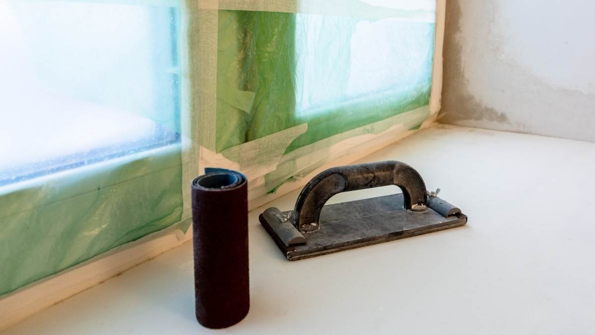 De binnenzijde van een raam dat wordt afgewerkt. Het raam is volledig afgeplakt: er zit een doorzichtige folie voor het raam, vastgeplakt met schilderstape. De diepe dagkanten worden afgewerkt met pleisterwerk. Op de vensterbank staan een rol zwart schuurpapier en een zwarte vlakspaan.