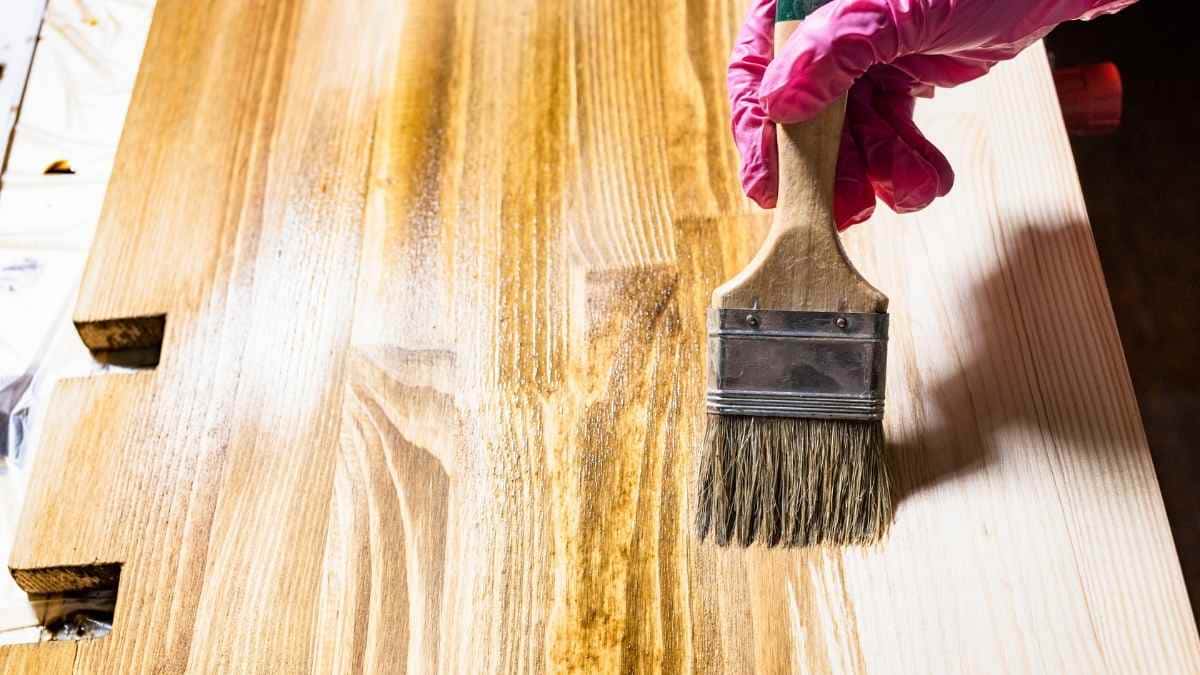 Een hand met roze handschoen gebruikt een kwast om een houten deur te vernissen. De deur bestaat uit licht houten planken. Recht in het deurblad zitten twee uitsparingen voor scharnieren.