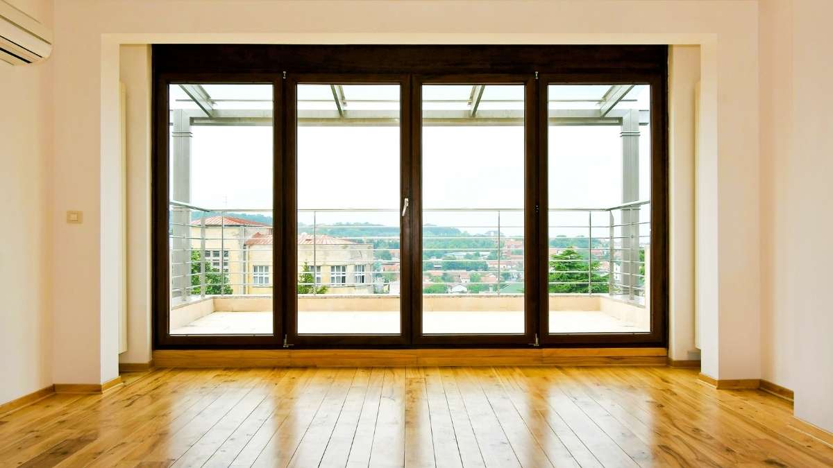 Een ruimte met een glanzend parket, witte muren en een dubbele glazen deur tussen 2 grote ramen. De deuren en de ramen zitten in een donkerbruin kozijn. Achter de dubbele deur ligt een terras met metalen balustrade en terrasoverkapping.