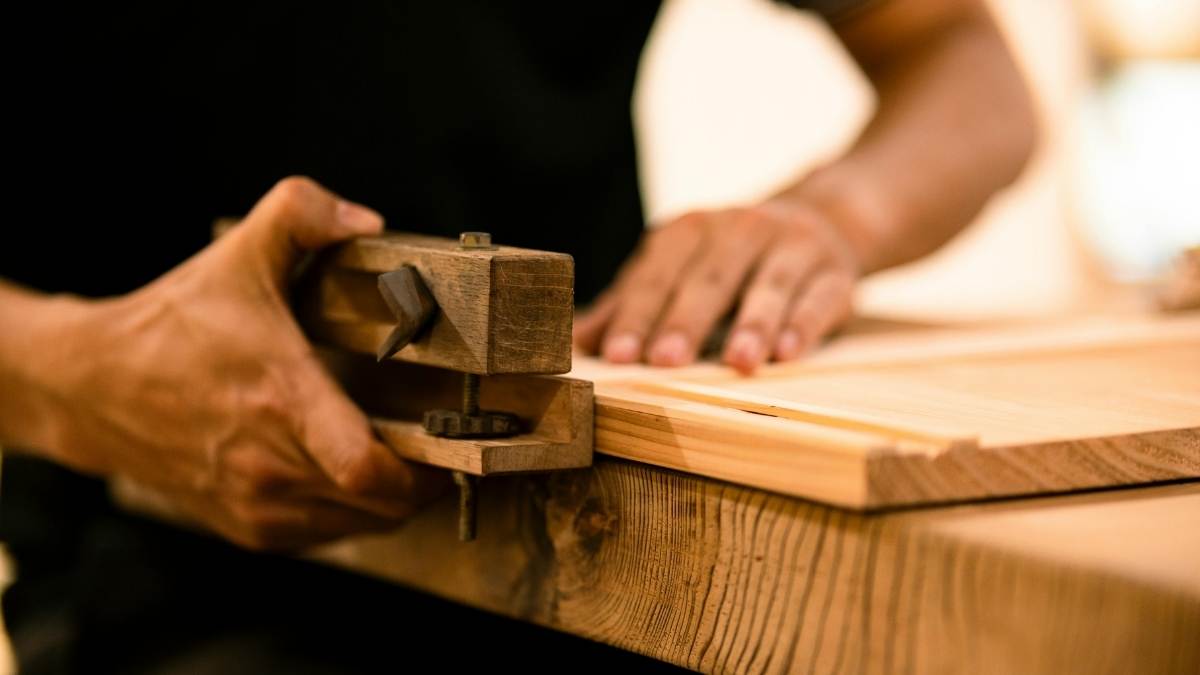 Een man gebruikt een manuele bovenfrees om een houten plank te bewerken. De plank bestaat uit licht gekleurd hout en ligt op een houten tafel. De man draagt een zwart T-shirt.