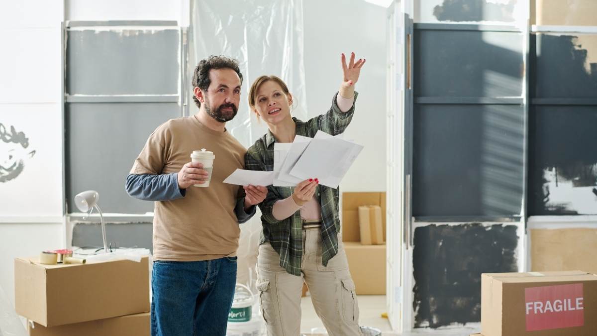 Een man in bruine trui en jeansbroek staat naast een vrouw in beige cargopants en groen geruit hemd. De man heeft een wegwerpbeker met koffie vast. De vrouw heeft papieren met renovatieplannen vast en wijst voor zich uit. De blik van de man volgt haar hand. Ze staan in een half afgewerkte ruimte.