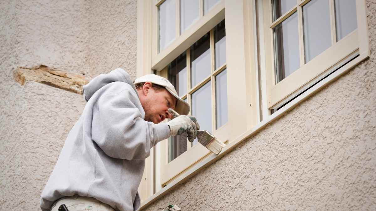 Een ramenspecialist in grijze trui en witte pet schildert de houten kozijnen van een dubbel raam. Hij gebruikt een gelige, crème-achtige kleur. Het raam zitten in een gevel die is afgewerkt met zandkleurig crepi.