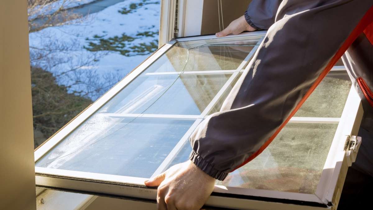 Een man verwijdert een raam met oud glas om het te vervangen. Enkel zijn armen en handen zijn in beeld. Hij draagt een grijze jas met oranje strepen. Het raam zit in een wit kozijn met roedes.