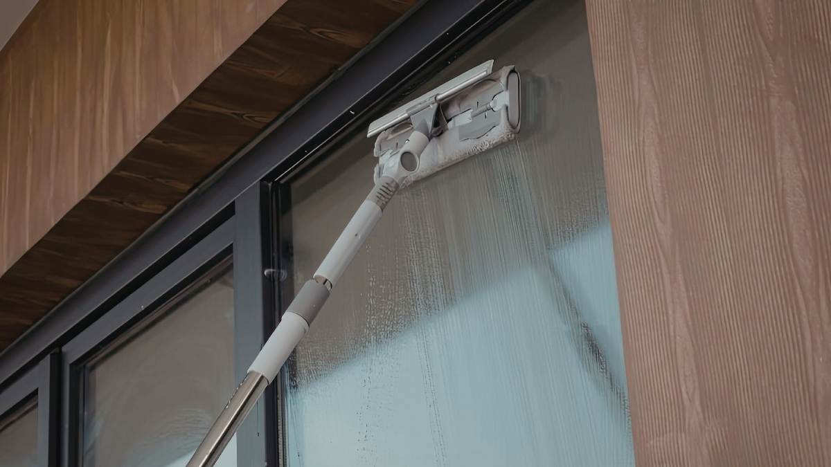 Een borstel op een telescoopsteel wordt gebruikt om hoge ramen met zwarte aluminium kozijnen te onderhouden. De ramen zitten in een donkerbruine houten gevel.