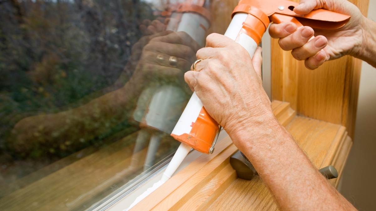 Close-up van twee handen die een oranje kitpistool vasthouden. De kitkoker is wit. Uit de spuitmond komt de kitrand, die tussen de beglazing en het houten raamkozijn gespoten wordt.