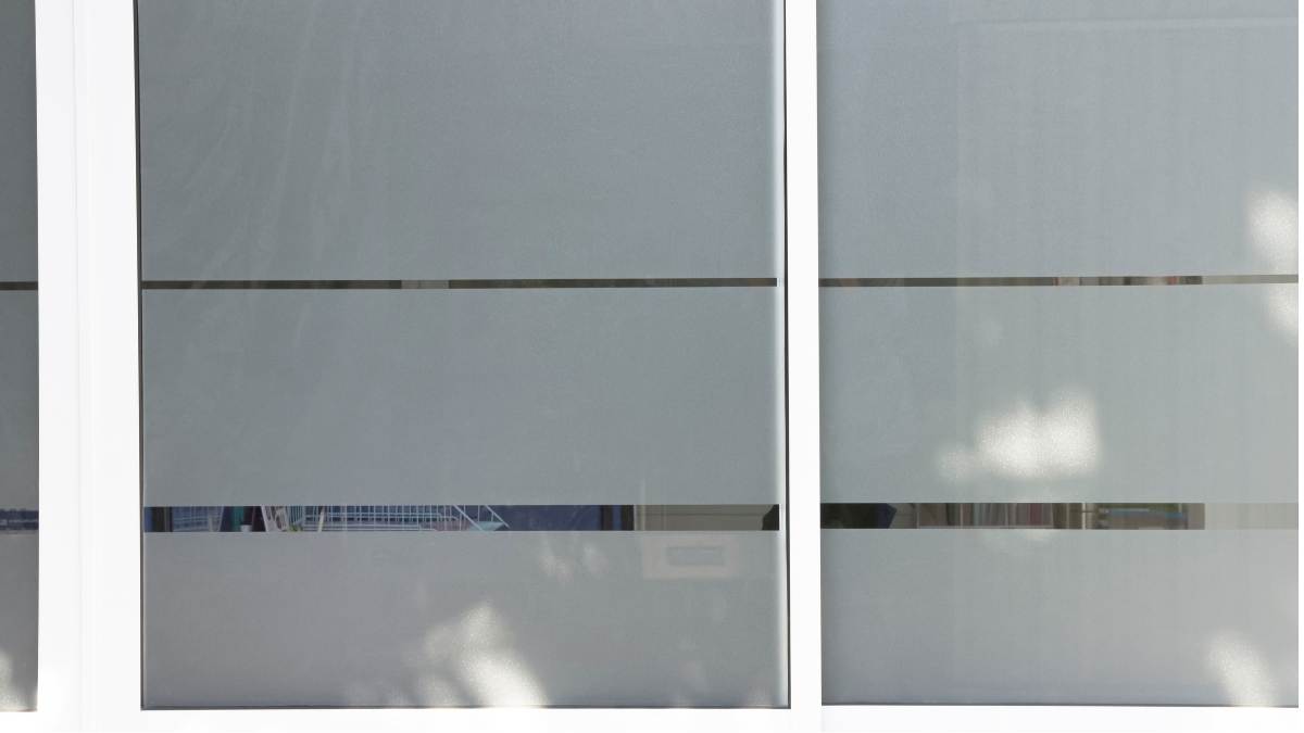 Een raam met witte kozijnen waarop een inkijkwerende privacyfolie is gekleefd. Er zitten twee smalle, horizontale insnijdingen in de folie. De bovenste insnijding is smaller dan de onderste.