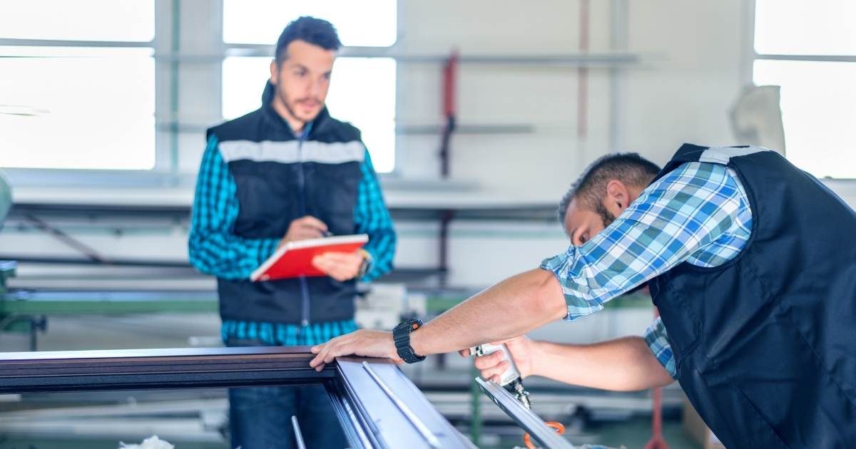 Een raamspecialist in een donkerblauwe bodwarmer over een lichtblauw met wit geruit hemd werkt aan een nieuw raam met driedubbel glas. Achter het raam staat een collega in dezelfde bodywarmer, met een iets donkerder blauw geruit hemd. De collega maakt notities in een rood notitieblok.