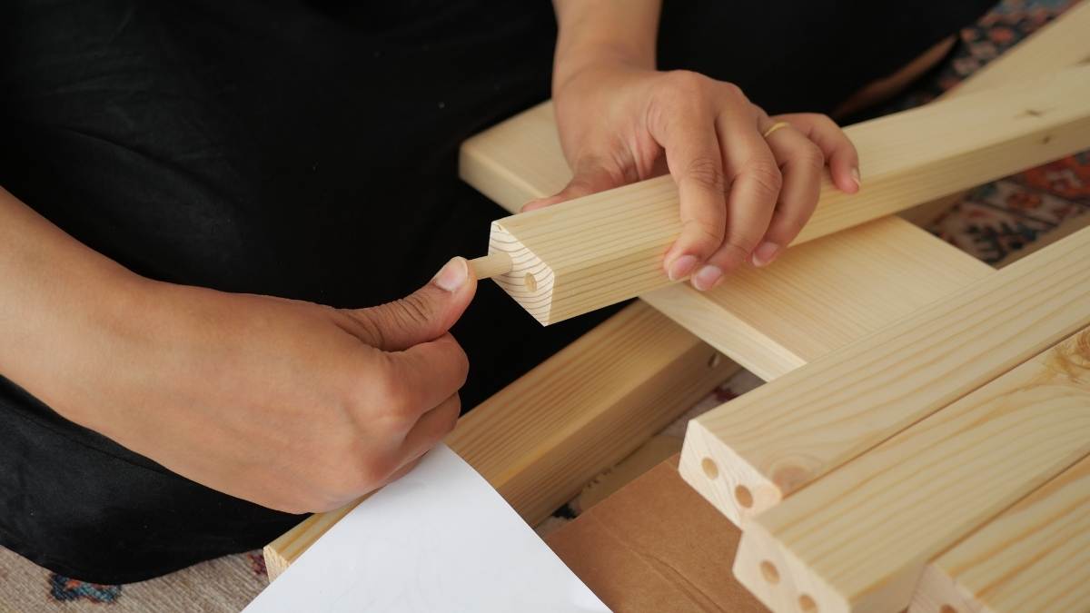 Twee handen hebben houten latten vast. De linkerhand houdt de lat vast, de rechterhand steekt een pin in de pen-en-gat-verbinding in de houten lat. Het hout is licht.