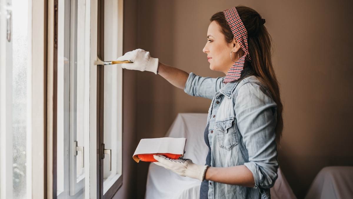 Een vrouw in een lichtblauw jeanshemd en een rood met blauw geruite hoofdband schildert de binnenzidje van haar kozijnen. Achter de vrouw staan meubels die zijn afgedekt met witte doeken. De muren van de ruimte hebben een diepbruine tint. Ze beschildert de ramen met zeer licht taupe of bruine verf.