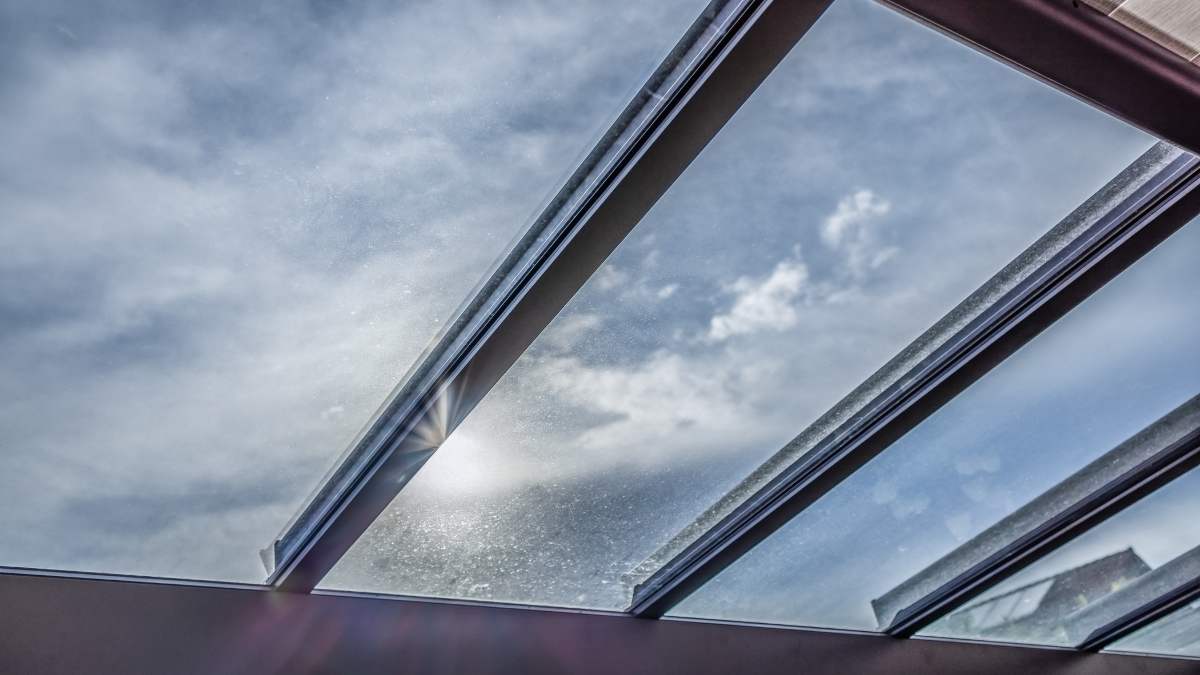 Het glazen dak van een veranda, voorzien van zonwerende 4-seizoensbeglazing. Boven het raam een bewolkte hemel, met een waterig zonnetje.